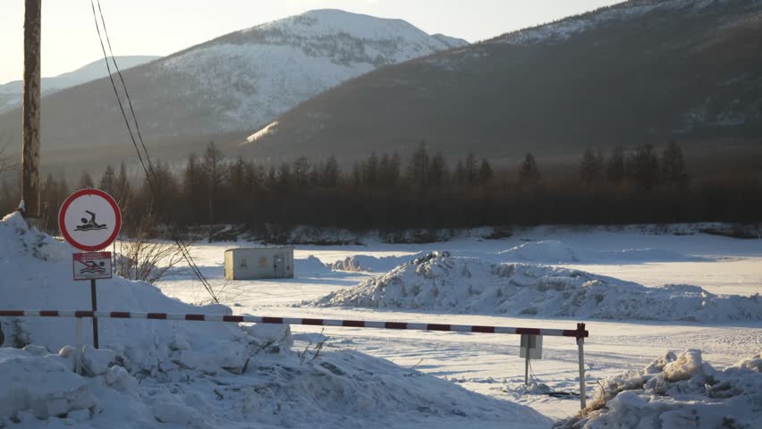 A snow-covered area shows a warning sign near a frozen lake. Mountains rise in the backdrop. The scene captures the cold winter day, showcasing nature and safety measures.