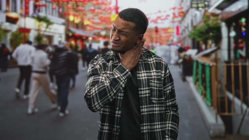 Young black man touching his neck and grimacing while standing on a crowded street market with lanterns and stalls; pain concern.