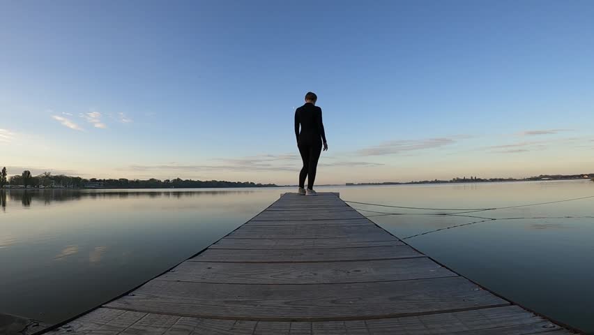 A woman in a black jumpsuit at dawn on a very beautiful blue background of a lake on a wooden walkway is engaged in sports, namely fitness, a woman does a warm-up