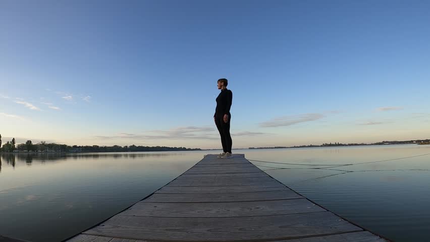 A woman walks along a wooden walkway after a warm-up, against the backdrop of a lake and the dawn sky.