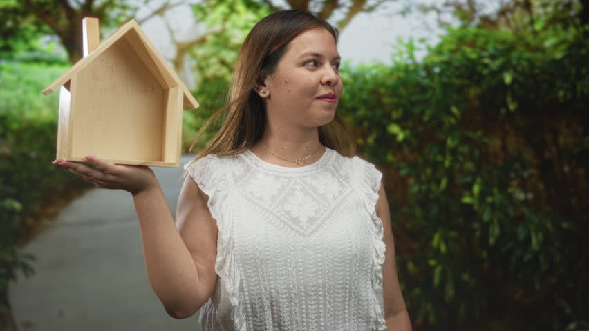 Young woman holding small wooden house on outstretched hand in forest walkway; hopeful homeownership ambition.