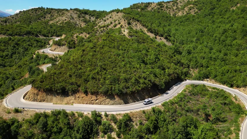 Aerial view of a campervan traveling, driving the iconic SH75 mountain road in Albania, winding through the rugged landscape above the vibrant turquoise waters of the Vjosa River in the summer sun.