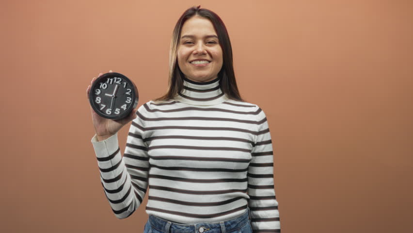 Woman in striped turtleneck holding a round clock with left hand and showing horns finger gesture with right bare hand in studio backdrop; joy celebration.
