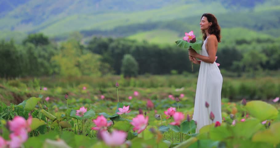 Wide shot of a woman in a white dress standing in a blooming lotus pond in Tra Ly, Quang Nam, Vietnam. Peaceful natural scenery with green hills. Ideal for travel, wellness, and nature content.