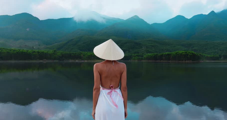 Wide shot of woman in white dress and conical hat walking along Tra Ly Lake in Quang Nam, Vietnam with mountain backdrop. Ideal for travel documentaries and tourism content.