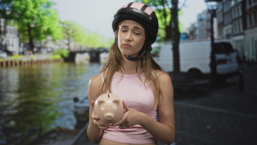 Young woman holding piggybank with helmet, showing bare shoulders on street in amsterdam; money worry.