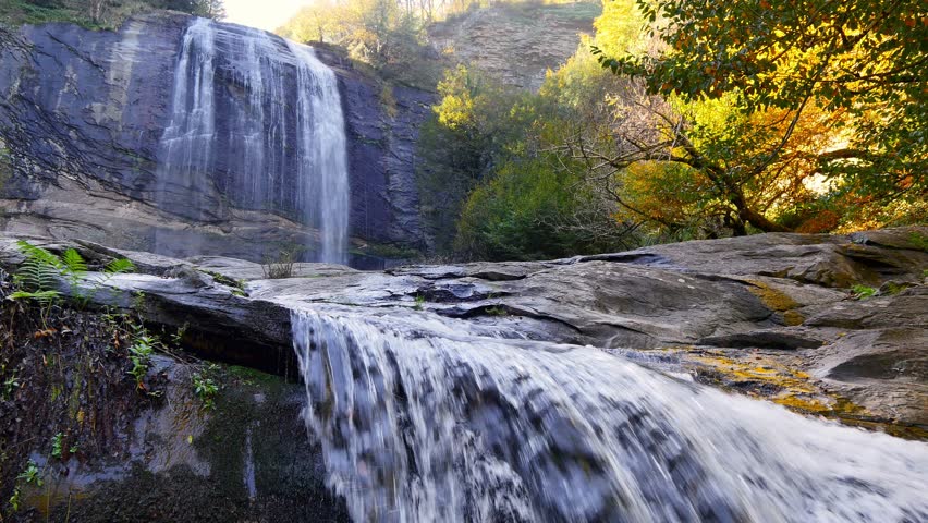 Waterfall cascading over layered rock ledges in autumn forest with flowing stream in foreground, sunlight illuminating colorful foliage and textured cliff in tranquil natural setting. Suuçtu, Bursa, Turkey