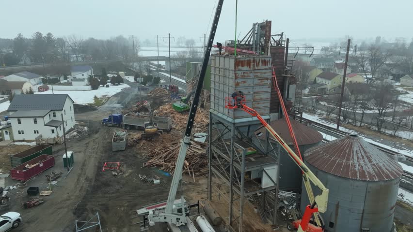 Workers are busy at a construction site where a new structure is being built. Cranes and vehicles move materials while homes are seen nearby. Snow covers the ground in this rural setting.