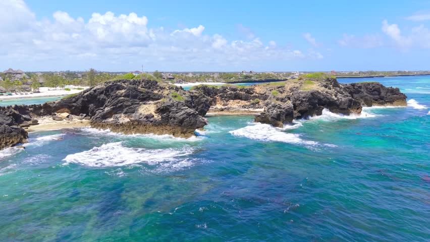 Aerial view of waves crashing against dark coral rock formations and turquoise water near a white sand beach in Watamu, Kenya.