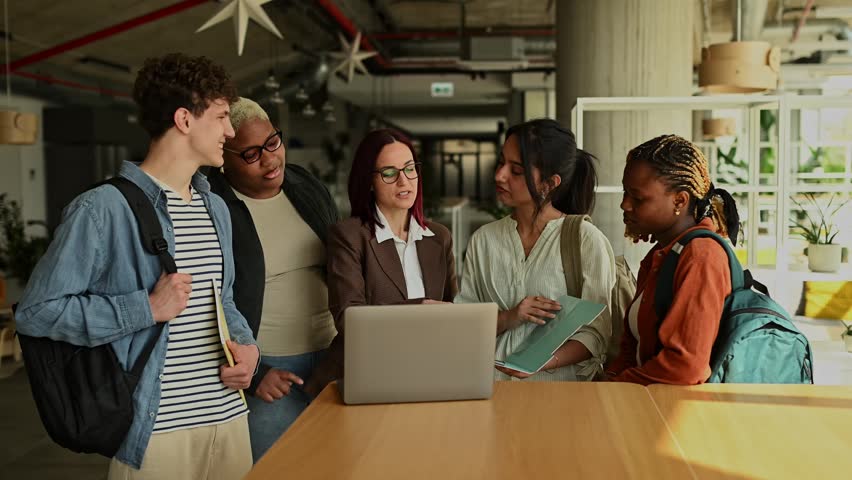 Diverse group of university students and their female professor collaborating on a project, gathering around a laptop in a campus common area