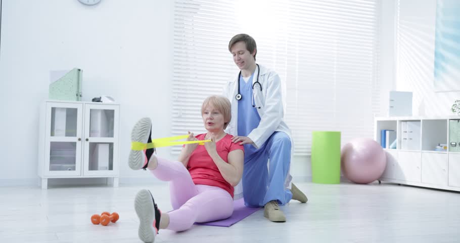 Senior woman training with resistance band under physiotherapist supervision indoors