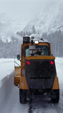 Yellow plow clearing mountain road in heavy snow, park maintenance operator drives across icy pathway flanked by towering granite cliffs and dense evergreen forest. Yosemite National Park