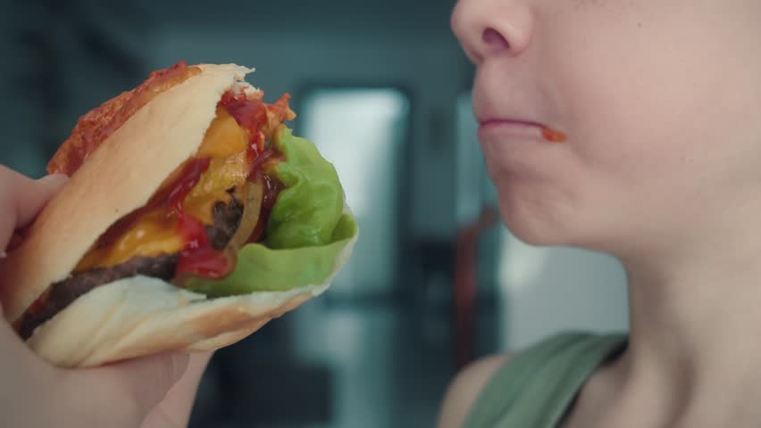 Child eating crispy chicken burger with lettuce, pickles, and tomato in restaurant. Close up shot of child biting hamburger. Hungry boy eating unhealthy food