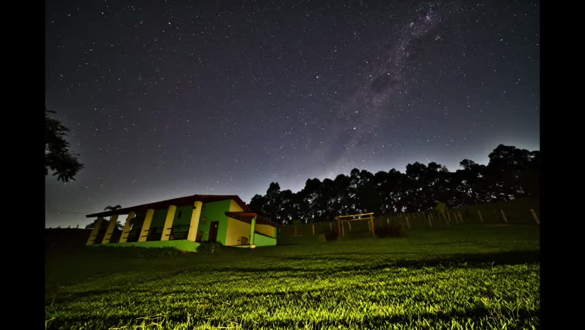 Night sky time-lapse of the Milky Way galaxy moving over a rural house and green pasture. Scenic view of the stars, celestial sphere and deep space nebula in a dark sky environment in the countryside.