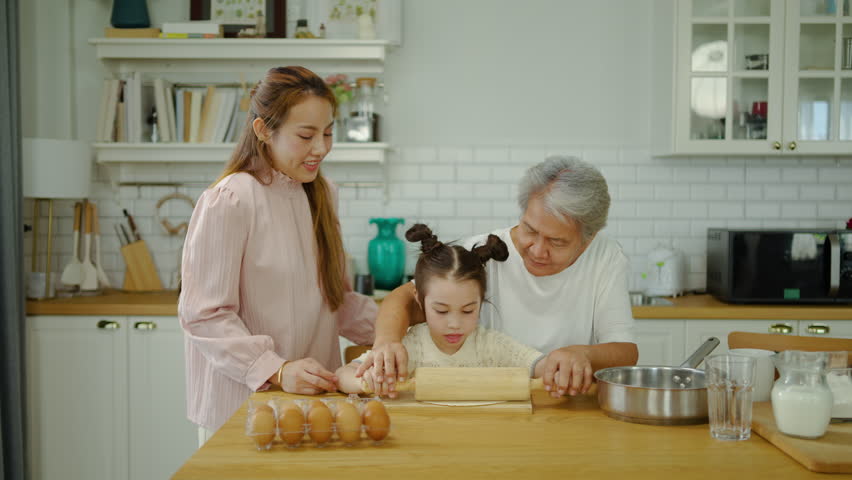 Happy Asian family with three generations baking together in a bright modern kitchen. Grandmother and mother teach a cute little girl how to use a rolling pin on dough. Joyful domestic lifestyle.