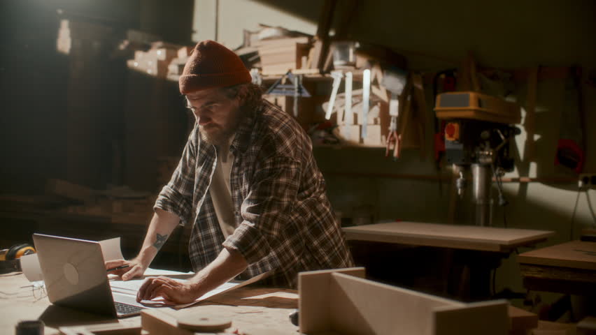 Craftsman using laptop and sketching plan on paper at workbench in carpentry workshop, preparing before woodworking project
