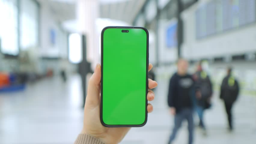 Hand holds phone with green screen. Background shows blurred airport terminal. People walk with luggage in distance. Phone frame visible around screen. Focus on phone, not surroundings
