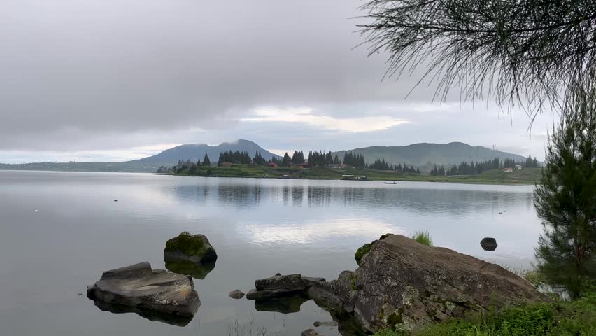 A video of a lake with calm waters surrounded by natural rocks, with mountains and hills in the background
