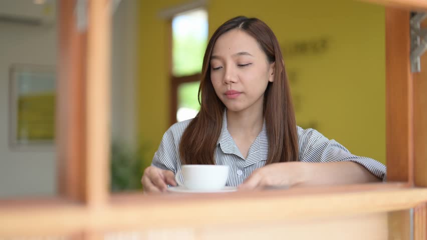 A woman is sitting at a table with a cup of coffee in front of her. She is looking at the camera with a serious expression