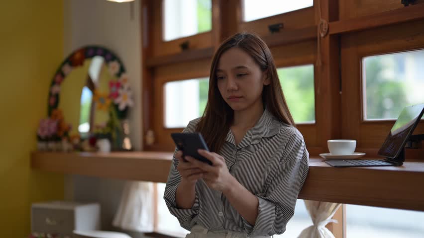 A woman is looking at her cell phone while standing in front of a window. She is focused on her phone and not paying attention to her surroundings