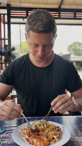 Close-up of a man eating Pad Thai noodles with shrimp at a local street restaurant in Thailand. Authentic Asian street food experience, travel lifestyle, casual dining and traditional Thai cuisine