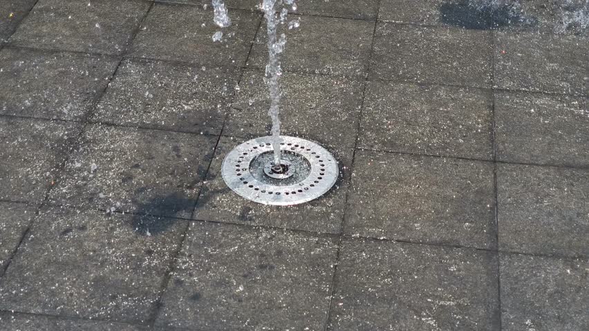 Water jet spraying upward from a ground fountain and splashing onto tiled pavement, captured in close up video, showing dynamic motion and splash detail for urban, lifestyle, and water feature visuals