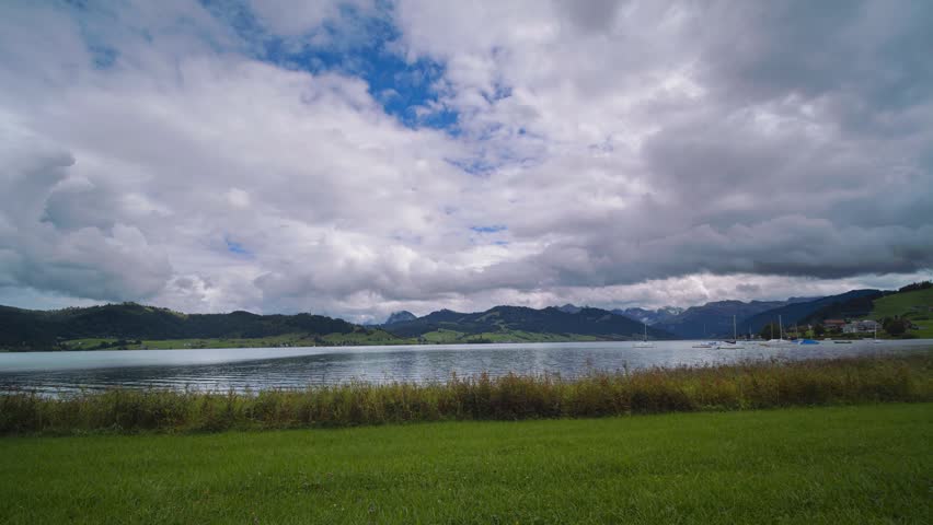 Wide panoramic view of a calm lake with mountains in the background under a cloudy sky.