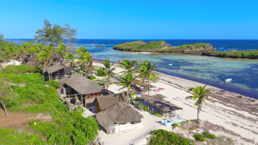 Aerial view of thatched roof huts and palm trees along the white sand coastline with rocky islands and clear water at Watamu Beach, Kenya.