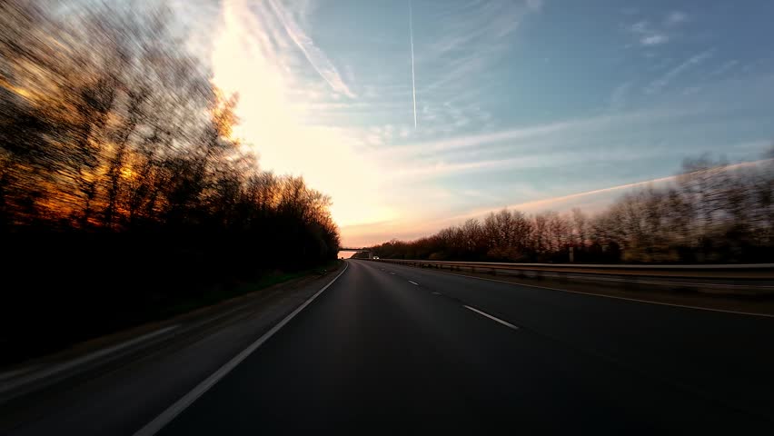 Truck driving along highway passing under bridge at sunset with trees, rural travel journey first person view. Straight asphalt road leading forward under overpass with evening sky glow, road trip