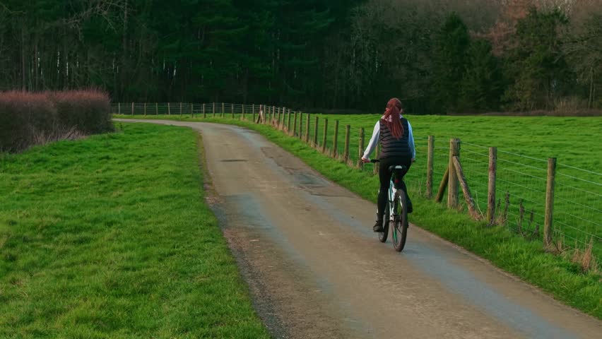 Female cyclist riding bicycle along winding countryside road between green fields and forest. Middle aged woman cycling rural lane promoting healthy lifestyle outdoor fitness and active leisure
