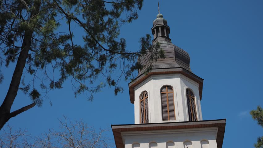 Low-angle travelling shot of a traditional church tower captured under a clear blue sky, framed by surrounding tree branches