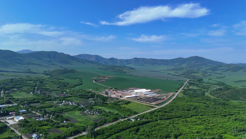 Drone footage captures a quaint small village with houses, roads, and paths nestled at the foothills of low mountains covered in green trees and meadows. Adjacent to the vast Bukhtarma Reservoir, the scene features lush vegetation, serene rural charm, and expansive water views under clear skies