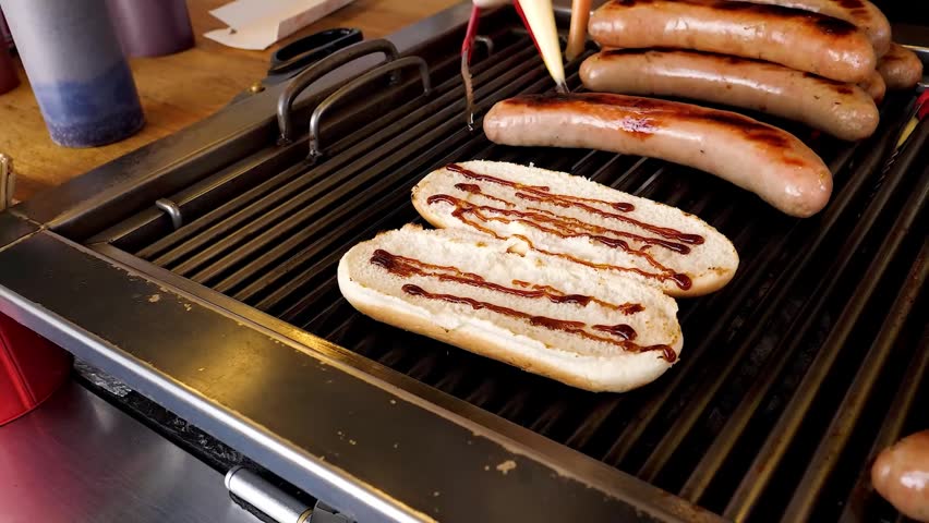 Close-up shot of a street vendor preparing hot dogs on a professional roller grill. Mustard and ketchup are being applied to buns, with sausages cooking in the background