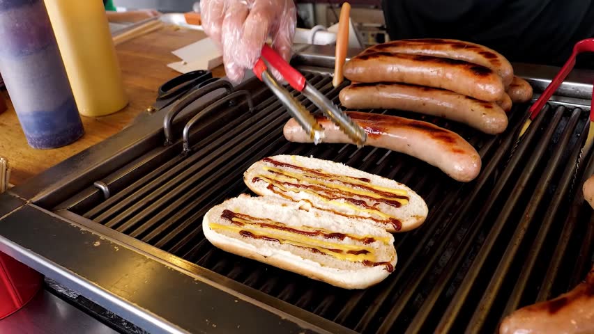 A close-up shot of several sausages and a single hot dog bun being grilled on a hot, professional-style barbecue grill. Tongs are visible in the background, and condiments are blurred in the foregroun