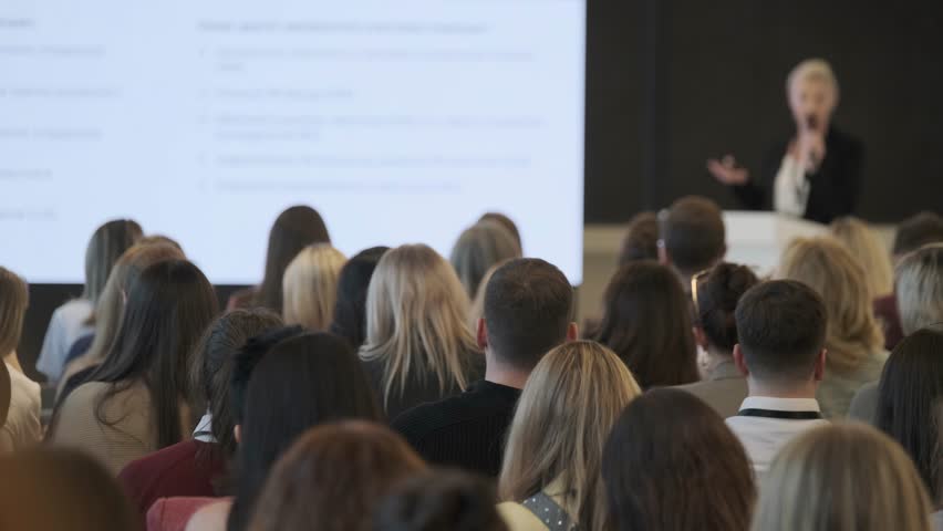 Large audience listening to presenter during business conference presentation, focus on attendees and professional learning environment.