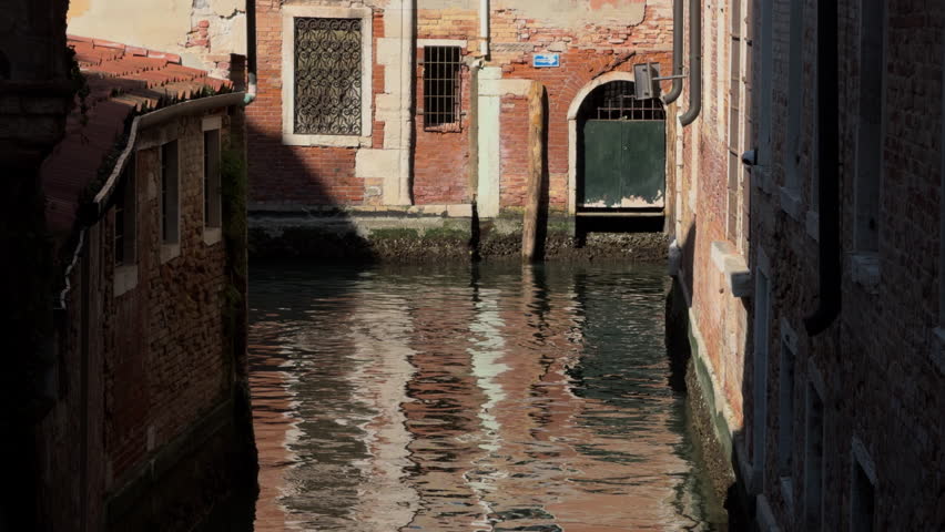 Slow-motion view of a tranquil Venetian canal between aged brick buildings, with sunlight gently rippling and reflecting across the calm water surface.