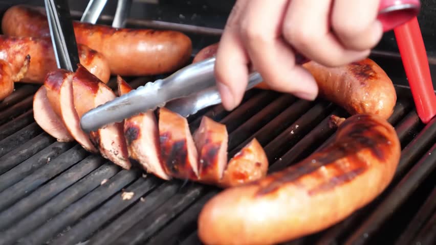 close-up shot of juicy, golden-brown sausages (kielbasa or bratwurst) grilling on a hot, cast-iron barbecue grill over charcoal. A hand uses metal tongs to adjust the sausages, one of which has cut ma
