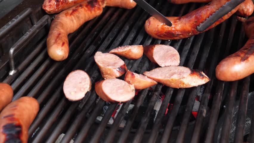 A close-up view of juicy, pre-cooked sausages sizzling on an outdoor charcoal grill. A person uses tongs and a knife to slice one of the sausages into pieces as they cook over the hot coals.