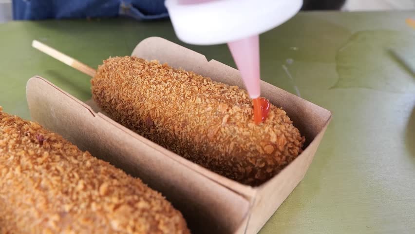 Close-up shot of a person squeezing a zigzag pattern of red ketchup onto a freshly fried, crispy Korean corn dog served in a brown paper tray. Another corn dog is visible in the foreground.