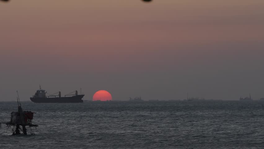 Red sun setting over ocean with distant industrial skyline and ships, atmospheric seascape for travel and environment themes