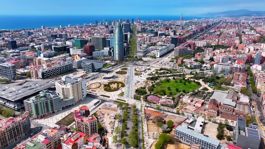 Aerial view of Plaça de les Glòries and Torre Glòries in Barcelona, Spain, showing modern architecture, broad boulevards, urban parks, and the Mediterranean coastline on a clear day.