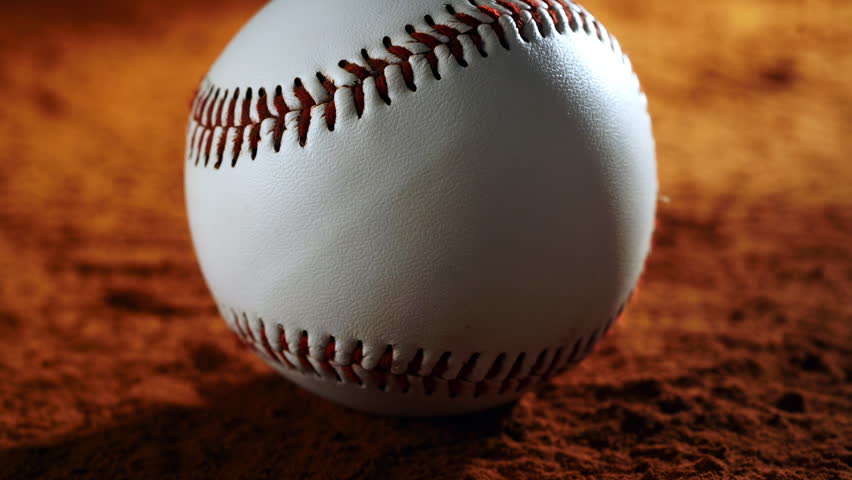 Cinematic close-up of a baseball resting on dusty ground as a gust of wind lifts dirt and particles around it. Dramatic lighting enhances texture and atmosphere.	