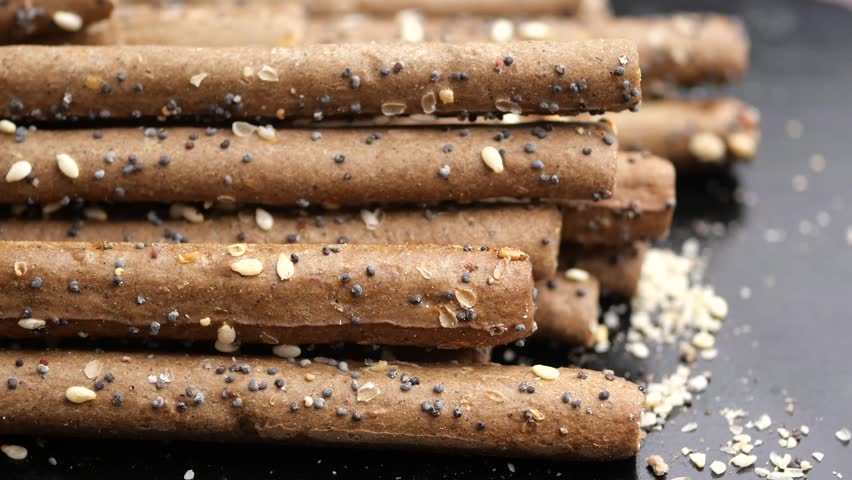 Dynamic motion footage of brown grissini breadsticks with poppy and sesame seeds in a stack, culinary texture detail.