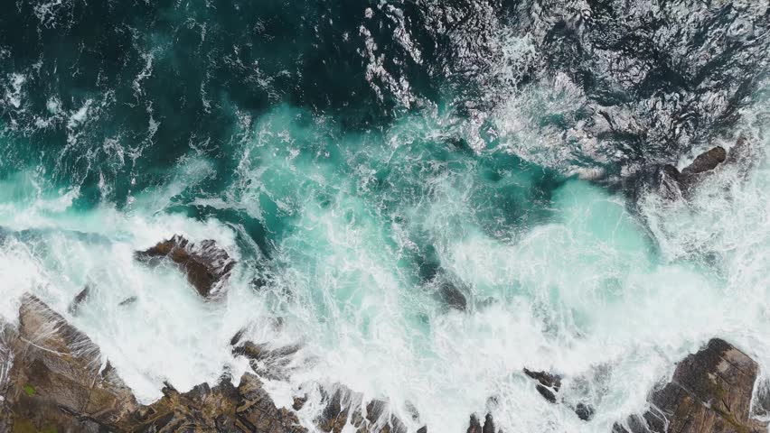 High-angle drone shot of turbulent ocean waves with white foam violently crashing onto rugged rocky coast in Nova Scotia, Canada. Dynamic water texture and constant motion during sunny day 4K