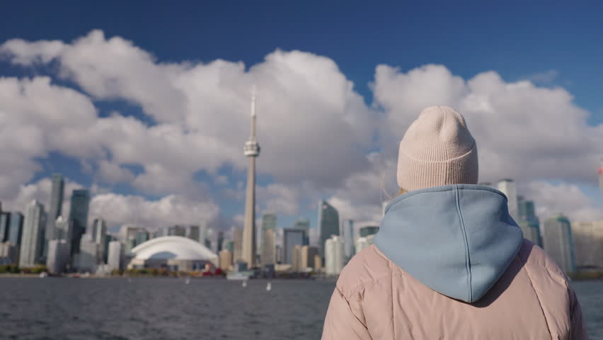 Young woman tourist looking at Toronto skyline from Lake Ontario waterfront, back view of teenager in warm jacket and hat enjoying urban cityscape and travel mood.