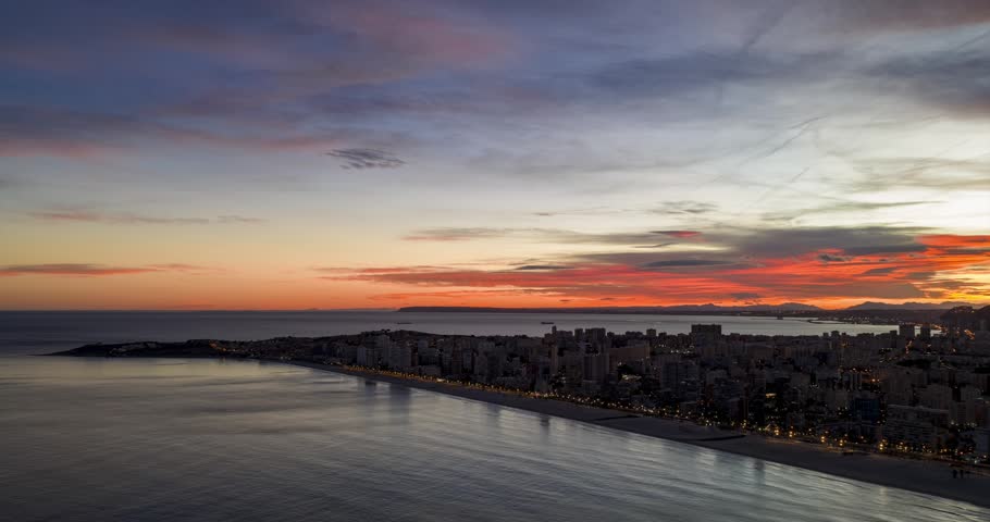 City lights along the coast turning off as a vibrant orange sun rises over the sea in Alicante, Spain. Aerial Hyperlapse, copy space