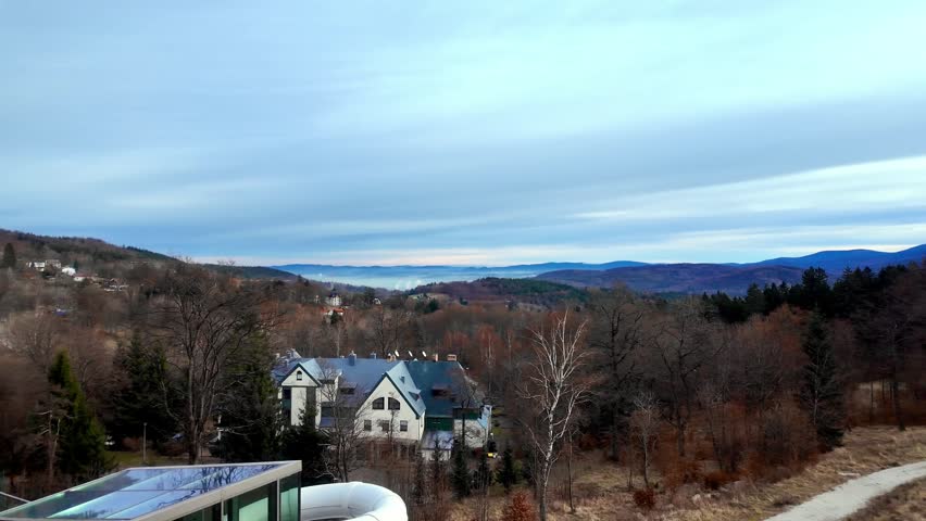 Picturesque landscape showing traditional houses, bare winter trees, and a dirt path, leading to misty mountains under a calm, cloudy sky