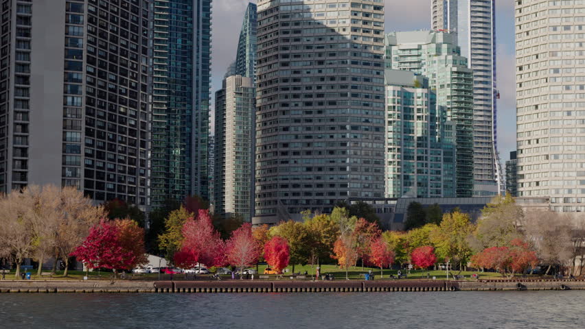 Toronto waterfront park with colorful autumn trees and modern high rise buildings, vibrant cityscape with red foliage along Lake Ontario shoreline.
