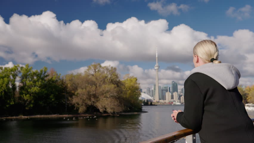 Young woman tourist leaning on boat railing looking at Toronto skyline across Lake Ontario, peaceful travel moment with autumn trees and calm water.