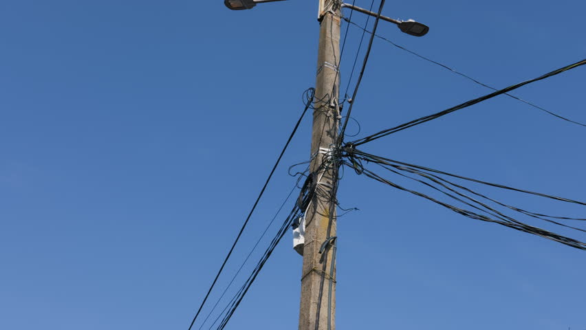 Concrete utility pole holds a chaotic tangle of black electrical and communication cables under a bright clear blue sky. The scene illustrates an urban infrastructure overload concept.